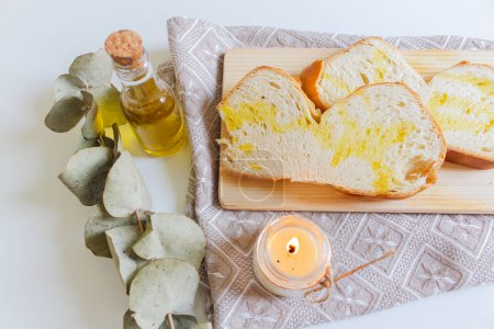 Foto de Plato mediterráneo con aceite de oliva y pan en la tabla de cortar. Acogedora decoración con vela ligera y hojas secas de eucalipto - Imagen libre de derechos