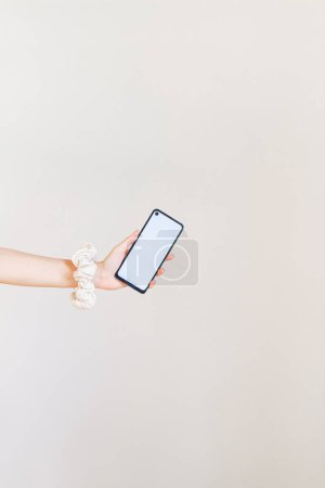 Téléchargez les photos : Main féminine avec scrunchie blanc au poignet tenant un smartphone à écran blanc - en image libre de droit