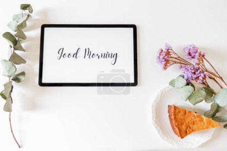 Foto de Frase "buenos días" en una tableta con flores secas, hojas de eucalipto y trozo de pastel sobre fondo blanco - Imagen libre de derechos