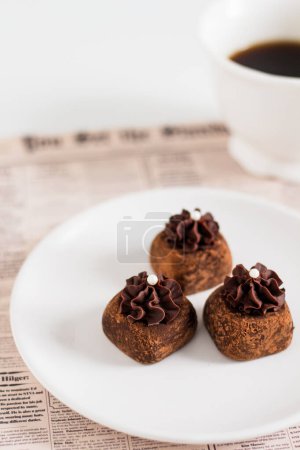 Téléchargez les photos : Bonbons gourmands au chocolat sur plaque blanche avec une tasse de café sur fond de journal. Stylisme alimentaire. - en image libre de droit
