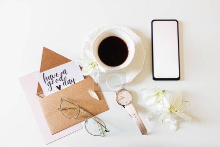 Photo for Inspirational quote: "have a good day" inside a kraft envelope. Home office morning composition with: mobile phone, wrist watch, white flowers, glasses and a cup of coffee. Flat lay, top view. - Royalty Free Image