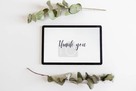 Téléchargez les photos : Phrase "Merci" sur une tablette numérique, cadre fabriqué à partir de feuilles d'eucalyptus sur fond blanc. Concept matin lent. - en image libre de droit