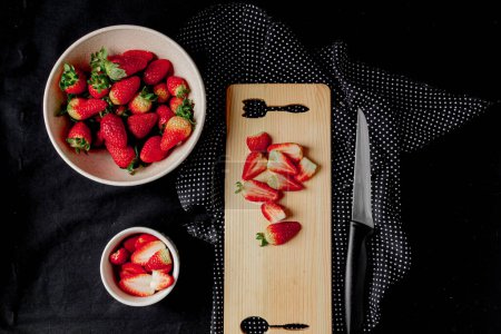 Photo for Top view of juicy strawberries in a bowls and some strawberries cut on the chopping board on a black background. - Royalty Free Image