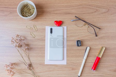 Photo for Home office desk mock up with smart phone, glasses, pen, sharpener, red lipstick, heart shape audio divider, headphone golden paper clips and wild flowers on wooden background. Flat lay, top view. - Royalty Free Image