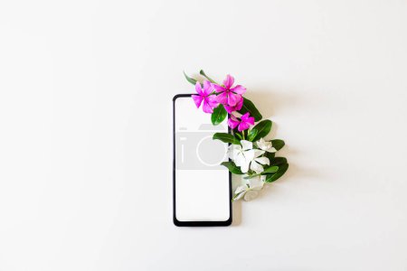 Photo for Smartphone with blank screen, copy space and purple and white flowers on white background. Flat lay, top view. Spring concept. - Royalty Free Image