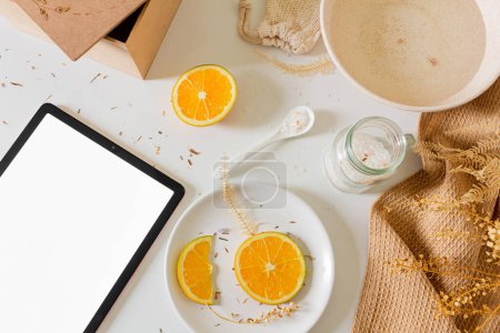 Photo for Beauty spa beige composition with wooden box, bowl with water, orange slices, blank screen tablet and bath salt on white background. Flat lay, top view. Female beauty treatment routine concept. - Royalty Free Image