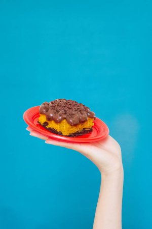 Téléchargez les photos : Main féminine tenant un morceau de gâteau aux carottes dans une assiette rouge sur fond turquoise. - en image libre de droit