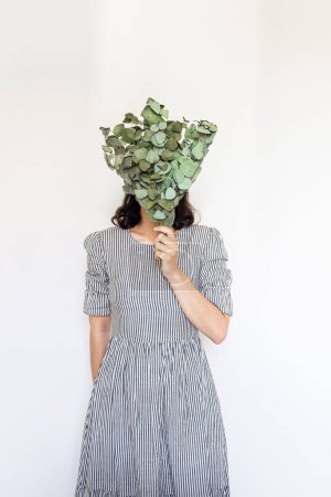 Téléchargez les photos : Jeune jolie femme en robe vintage rayée tenant un bouquet d'eucalyptus sur fond blanc. Fleuriste concept minimal. - en image libre de droit