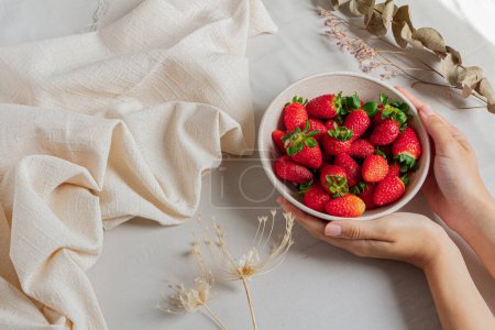 Téléchargez les photos : Jeunes mains féminines tenant un bol rempli de fraises sur un fond clair stylisé. - en image libre de droit