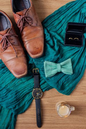 Foto de Accesorios elegantes de los hombres. Zapatos marrones sobre fondo de madera y tela verde. Vista superior de corbata de lazo, anillos de oro de la boda y un reloj de pulsera. Preparación para un concepto de boda. - Imagen libre de derechos