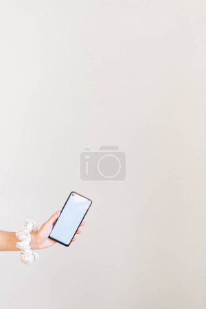 Foto de Mano femenina con scrunchie blanco en la muñeca sosteniendo un teléfono inteligente de pantalla en blanco - Imagen libre de derechos