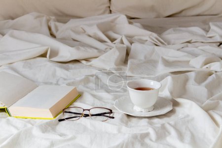 Photo for Open book with glasses and cup of tea on bed sheet - Royalty Free Image