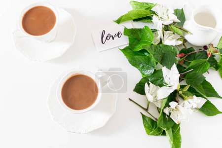 Téléchargez les photos : Tasses à lait et café, message d'amour sur une carte en papier avec des fleurs blanches et des feuilles de mûrier sur fond blanc. - en image libre de droit