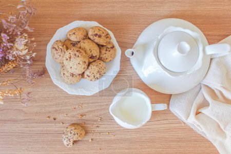Téléchargez les photos : Composition petit déjeuner. pile de biscuits dans une assiette blanche avec théière, pot de lait et bouquet de fleurs séchées sur table en bois - en image libre de droit