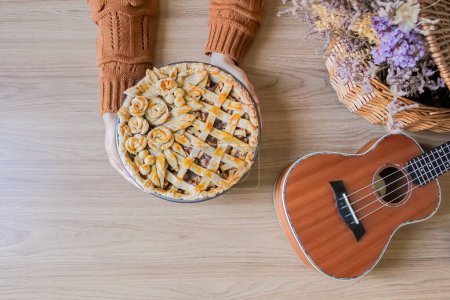 Foto de Composición otoñal con flores secas en cesta de picnic, ukelele y mujer joven poniendo pastel de manzana decorado - Imagen libre de derechos