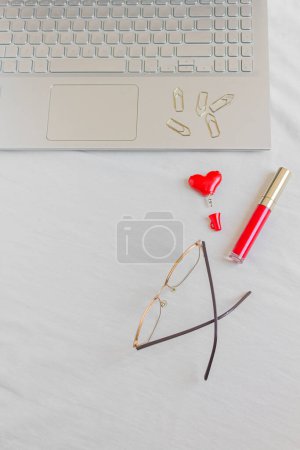 Foto de Espacio de trabajo de escritorio de oficina en casa con computadora portátil, gafas, lápiz labial rojo, clips de papel y divisor de audio del corazón sobre fondo blanco. - Imagen libre de derechos
