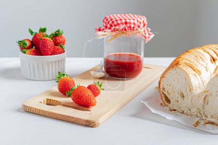 Photo for White linen fabric background with a jar of strawberry marmalade, a bowl full of fresh strawberries and a delicious bread. - Royalty Free Image