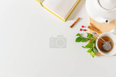 Foto de Taza de composición de té con colador de té, tetera, libro abierto, canela, hojas verdes plantas sobre fondo blanco. Piso tendido, vista superior. - Imagen libre de derechos