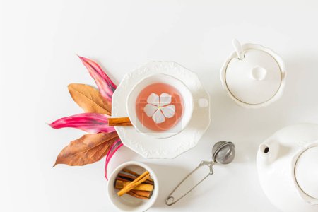 Foto de Hora del té composición rosa pálido con una taza de té con una flor blanca en el interior, hojas rosadas y marrones, tetera, canela sobre fondo blanco. - Imagen libre de derechos