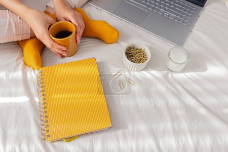 Foto de Una mujer con pijama, una taza de café en una cama, con un planificador, algunos clips y cintas washi. Freelancer, trabajo desde casa, día perezoso, concepto de hogar acogedor. - Imagen libre de derechos