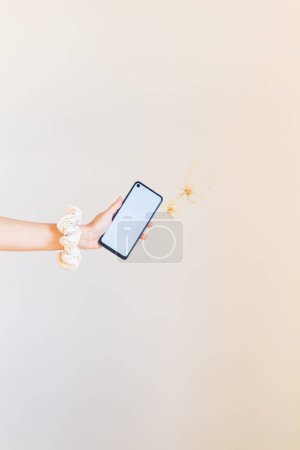 Foto de Joven mano femenina con scrunchie blanco en la muñeca sosteniendo un teléfono celular de pantalla en blanco y algunas flores secas - Imagen libre de derechos
