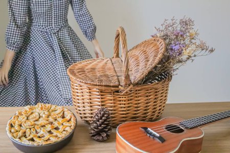 Foto de Composición otoñal con flores secas en cesta de picnic, ukelele, tarta de manzana decorada y mujer joven de pie sobre el fondo - Imagen libre de derechos