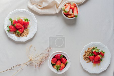 Téléchargez les photos : Fraises sur une assiette de granola et de miel, fraises également dans un bol et un sucrier. Tissu à pois noir et blanc. Fond blanc. - en image libre de droit