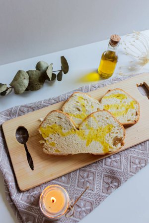 Foto de Plato mediterráneo con aceite de oliva y pan en la tabla de cortar. Acogedora decoración con vela ligera y hojas secas de eucalipto - Imagen libre de derechos