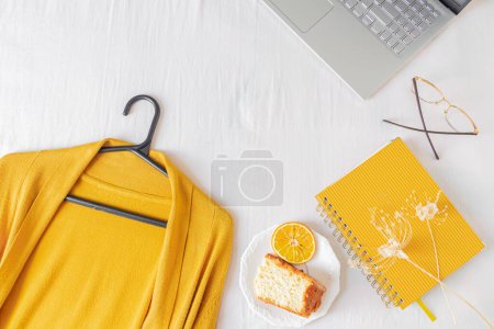 Téléchargez les photos : Bureau à domicile cadre avec ordinateur portable, morceau de gâteau orange, cardigan, lunettes et planificateur sur fond blanc. - en image libre de droit