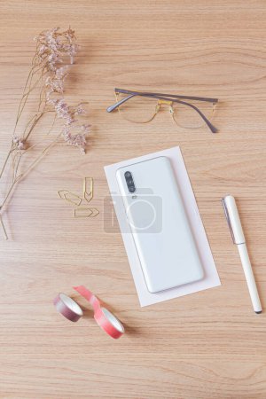Photo for Home office desk mock up with smart phone, glasses, pen, sharpener, golden paper clips and wild flowers on wooden background. Flat lay, top view. Lady boss concept. - Royalty Free Image