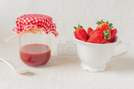 Téléchargez les photos : Marmelade de fraises sur un pot et pleine tasse de fraises fraîches sur un fond blanc. Concept de repas saisonnier. - en image libre de droit
