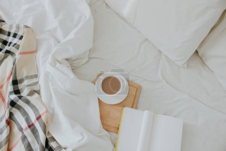 Photo for Cup of coffee on wooden board in bed with book and light beige sheet and pillows. Flat lay, top view. Slow romantic morning concept. - Royalty Free Image