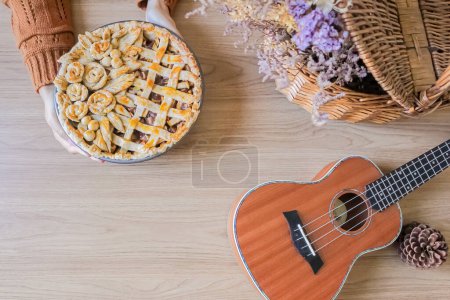 Téléchargez les photos : Mains féminines mettre décoré tarte aux pommes sur une table avec des fleurs séchées et ukulélé. Composition d'automne - en image libre de droit