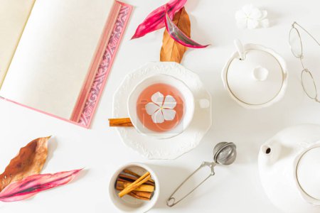 Foto de Hora del té composición rosa pálido con una taza de té con una flor blanca en el interior, hojas de color rosa y marrón, tetera, canela y libro abierto sobre fondo blanco. - Imagen libre de derechos