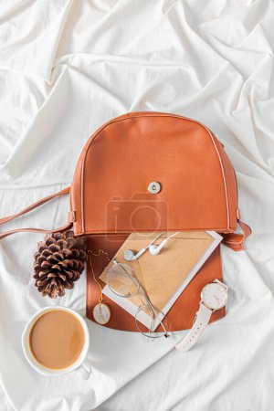 Photo for Fashion composition with female accessories and stationary on messy bed background. Bag with craft envelopes, earphones, glasses, necklace, wrist watch. Flat lay, top view. Autumn, fall concept. - Royalty Free Image
