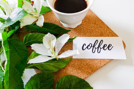 Photo for Black coffee cup on cork board, white flowers, card with the phrase: "coffee", and blackberry tree leaves on white background. Flat lay, Top view. Spring concept. - Royalty Free Image