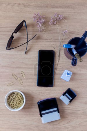 Photo for Mobile phone, stamps, heart paper cutter, paper clips, sunglasses and pens on wooden background. - Royalty Free Image
