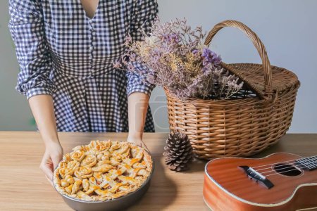 Téléchargez les photos : Composition automnale avec fleurs séchées en panier pique-nique et ukulélé. jeune femme mettant maison tarte aux pommes sur la table - en image libre de droit
