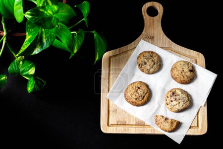 Photo for Cookies on wooden cutting board, boa plant leaves on black background. Close up. - Royalty Free Image