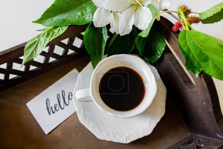 Téléchargez les photos : Tasse à café noire à l'intérieur d'un plateau, carte avec le message : "bonjour", fleurs blanches et feuilles de mûrier sur fond blanc. Concept de printemps. - en image libre de droit