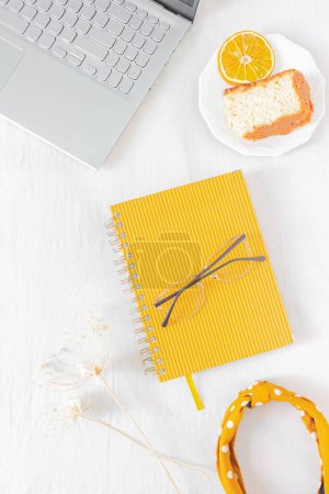 Photo for Home office desk frame with laptop, orange piece of cake, headband, glasses, beige wildflowers and notebook on white background. Flat lay, top view. Feminine business concept. - Royalty Free Image