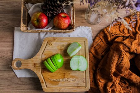 Photo for Autumn composition with sliced green apples on wooden board, straw basket with red apples and pine cone and brown sweater - Royalty Free Image