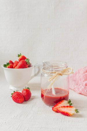 Téléchargez les photos : Marmelade de fraises sur un pot, une petite cuillère blanche à l'intérieur et des fraises fraîches sur un bol. Fond clair. Concept de repas saisonnier. - en image libre de droit