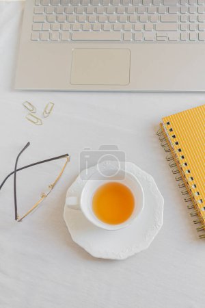 Photo for Home office desk workspace with laptop, cup of tea, glasses, paper clips and notebook on white background. Flat lay, top view. Women's, girl boss work or business concept. - Royalty Free Image