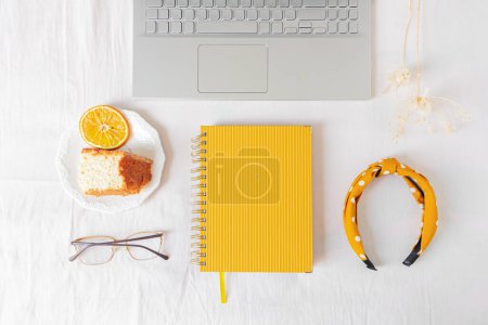 Téléchargez les photos : Bureau à domicile cadre avec ordinateur portable, morceau de gâteau orange, planificateur, verres et fleurs séchées sur fond blanc. - en image libre de droit