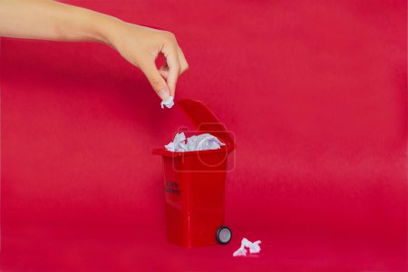 Téléchargez les photos : Main féminine mettant du papier à l'intérieur de la poubelle sur fond rouge. Concept de recyclage. - en image libre de droit