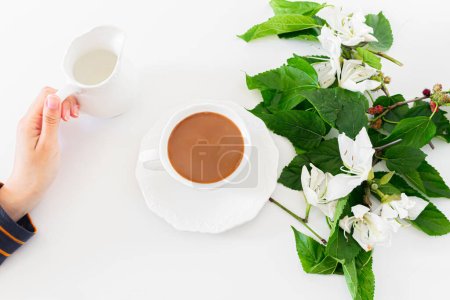 Photo for Female hand holding a jar of milk. Cup of coffee and milk, white flowers and blackberry tree leaves on white background. Flat lay, Top view. Spring concept. - Royalty Free Image