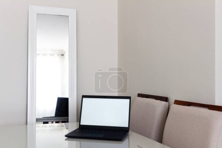 Photo for Laptop with copy space on the screen on the table. Stylish beige color dinner room with table, chair and curtains reflection in the mirror. Modern classic interior design. Comfortable home. - Royalty Free Image
