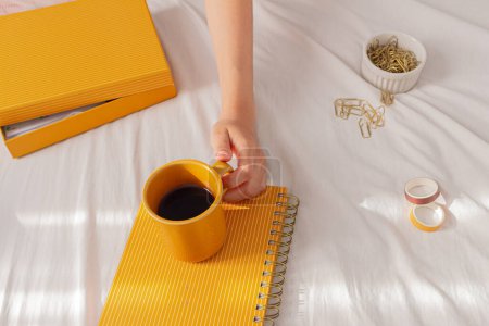 Foto de Una mujer con pijama, una taza de café en una cama, con un planificador, algunos clips y cintas washi. Freelancer, trabajo desde casa, día perezoso, concepto de hogar acogedor. - Imagen libre de derechos