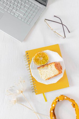Photo for Home office desk frame with laptop, orange piece of cake, headband, glasses, beige wildflowers and notebook on white background. Flat lay, top view. Feminine business concept. - Royalty Free Image